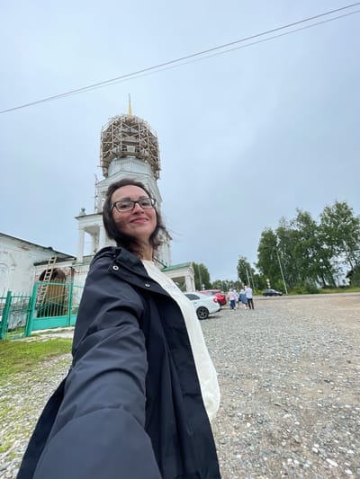 In the city of Kstining, Russia, a woman with glasses and dark hair is taking a selfie in front of a white church. The church has a dome that is currently under scaffolding, suggesting it is undergoing renovation. To the left of the church, there is a green fence and some grassy areas. In the background, several cars are parked, and a group of people are walking together. The sky is overcast. The ground in the foreground is covered with gravel. The woman is wearing a black jacket and a white shirt.