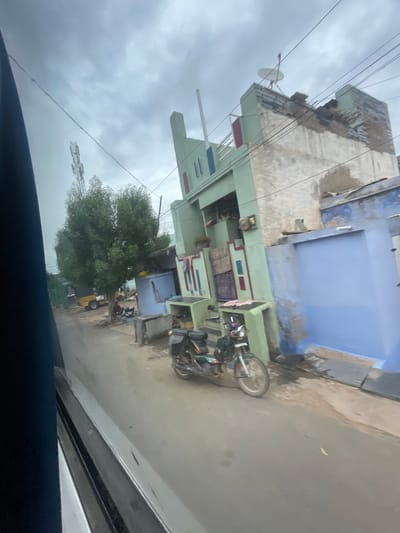 The image depicts a street scene in Bethamcherla, India, under an overcast sky. A multi-story building, partially painted in light green and off-white, dominates the right side of the frame. The building has a satellite dish on its roof and appears to be in a state of disrepair in some sections. Adjacent to it is another building with a bright blue facade. In the foreground, a green and black motorcycle is parked on the paved street. Further down the street, a small yellow vehicle and trees are visible. There are also several utility poles and wires crisscrossing the sky. The overall impression is of a typical street in a semi-urban or rural Indian setting.