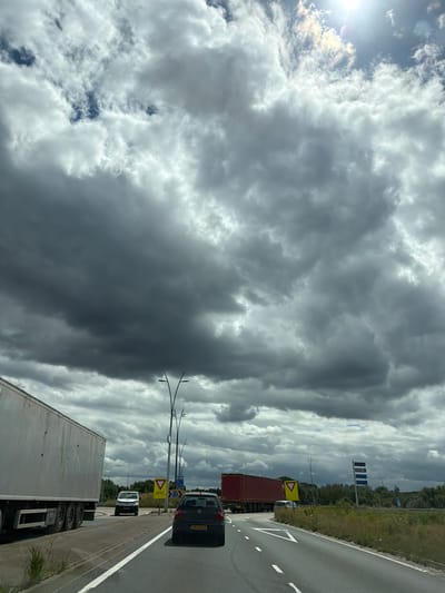 The image captures a view from a vehicle on a highway with a dramatic cloudy sky overhead. In the foreground, a white semi-trailer truck is on the left, and a black Volkswagen Polo with license plate "29-LH-LG" is in the right lane. Further ahead, a red truck and a white van are visible. Yellow traffic signs, including a yield sign, are present on the side of the road. The environment appears to be a rural or semi-rural area with trees and vegetation visible. The scene suggests a journey on a highway in or near Son en Breugel, Netherlands, given the presence of typical Dutch road signage and the overall landscape. The lighting indicates it is daytime, but the heavy cloud cover creates a somber atmosphere.