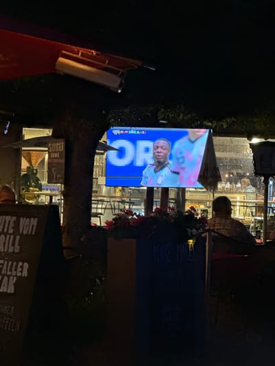 The image depicts an outdoor seating area at night, likely at a restaurant or beer garden in Prien am Chiemsee, Germany. A large screen displays a soccer match with players in light blue and pink uniforms. One player in the foreground is in focus, with the team name "FUSSBALL.TV" visible. The screen is illuminated by its own light, casting a blue glow on the surroundings. In the foreground, there are dark chalkboards with handwritten text, one of which appears to be a menu. A person is seated at a table in the lower right, facing away from the camera, watching the screen. Another person is visible through a window in the background, behind the screen. The environment includes plants, outdoor furniture, and architectural elements typical of a Bavarian establishment. The lighting is dim, with artificial lights illuminating the screen and some areas of the seating.