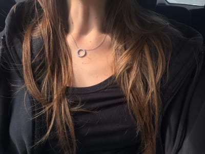 The image shows a close-up, overhead view of a person's neck and chest, framed by their long, brown hair. The person is wearing a black, ribbed garment and a black zip-up hoodie. A delicate silver necklace with a circular, diamond-encrusted pendant rests on their décolletage. The lighting suggests it is daytime, with a warm, directional light casting shadows and highlighting the textures of the hair and skin. A faint mark, possibly a hickey or a temporary stain, is visible on the upper chest. The background is dark and out of focus, with some suggestion of an indoor setting. There is no visible text in the image. The overall impression is intimate and casual, focusing on the jewelry and the soft interplay of light and shadow on the skin and hair. The location context of Navazi, Georgia, does not provide any specific visual cues within the image itself.