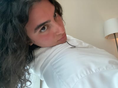 A woman with dark, curly hair is positioned on her side, resting her head against what appears to be a white surface, possibly a pillow or part of a white shirt. Her eyes are directed toward the camera. The background is a plain, light-colored wall with a lamp visible in the upper right corner. There is nothing that indicates this is El Caracol, Mexico.