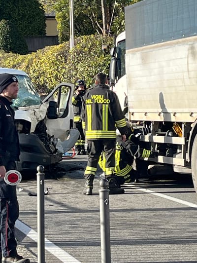 An emergency response scene is depicted on an asphalt road in Griante, Italy, following a collision between two vehicles. In the foreground, on the left, a male individual wearing a dark beanie and dark uniform with red side stripes holds a circular red-and-white traffic control paddle. To his right, two dark grey bollards stand upright on the road, which features faint white markings.
Multiple firefighters, identifiable by their dark uniforms with yellow reflective stripes and the text "VIGILI DEL FUOCO" on their backs, are actively engaged at the scene. One firefighter stands prominently in the center-right, facing away from the viewer. Another firefighter is visible further back near a severely damaged white van, whose front end is crushed and open. A third firefighter is crouched or kneeling on the right side of the image, appearing to work near the underside of a large white truck, which is partially visible and also involved in the incident. Red and blue hoses or cables are visible on the ground near the damaged van. The background features dense green foliage and trees under clear daylight conditions.