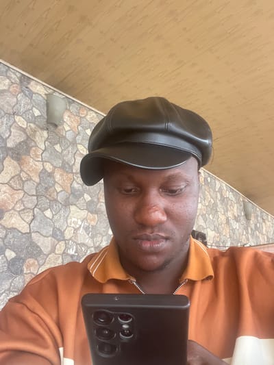 A person is holding a black smartphone with multiple camera lenses, possibly taking a selfie. The person is wearing a black leather hat and an orange sweater with white stripes at the collar. A thin silver chain is visible around the person's neck. The background includes a wall with a light brown, textured surface and a stone-tiled floor. The photograph was taken outdoors in the city of Nibo, Nigeria.