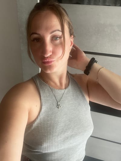 A woman is shown from the chest up, lying down and looking at the camera. She has light skin and dark brown hair pulled back from her face. She is wearing a grey, ribbed tank top and a delicate silver necklace with a star-shaped pendant. On her left wrist, she wears a black fitness tracker and a thin beaded bracelet. Her right hand rests on her head, near her ear. The background appears to be a textured, grey wall with vertical panels. The lighting suggests an indoor setting, possibly during the daytime. The woman's expression is neutral. There is no visible text in the image. The location context is Zarichne, Ukraine.