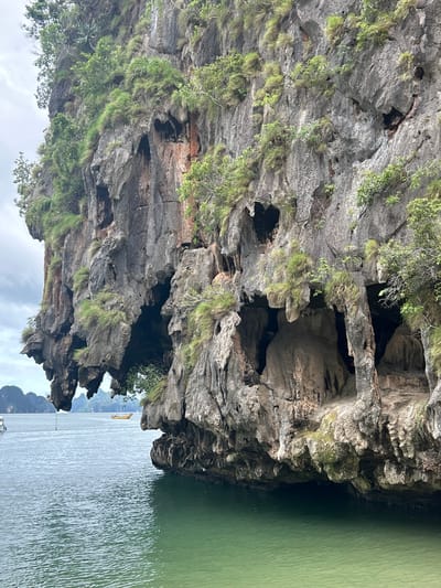 The image displays a towering limestone karst formation with lush green vegetation clinging to its rocky surface. The formation features numerous caves and overhangs, carved by erosion. The foreground is dominated by calm, turquoise-green water, reflecting the light and the submerged base of the karst. In the distance, a yellow boat is visible on the water, and the hazy outlines of other karst islands can be seen on the horizon under a cloudy sky. The scene suggests a tropical coastal environment, characteristic of areas like Tambon Krasom in Thailand, known for its dramatic limestone karsts and island formations. There are no people or specific human actions depicted, only the natural landscape and a distant boat.