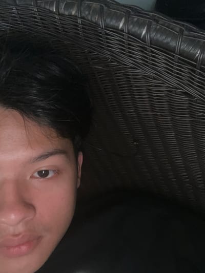 The image is a close-up, low-angle shot of a person's face, with the left side of their face and eye visible. The person has dark hair and brown eyes. The background consists of a dark, textured surface that appears to be wicker or rattan. The lighting is dim, casting shadows and highlighting the contours of the person's face. There are no indications of the specific location within Serdang Kulon, Indonesia.