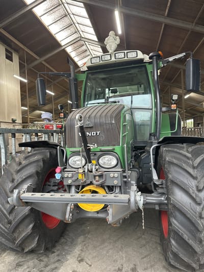 The image shows a close-up, head-on view of a green Fendt tractor parked inside a barn or agricultural building. The tractor is the primary subject, occupying most of the frame. It is a large, powerful machine with prominent tires. The building's interior is visible, characterized by a wooden roof with skylights and fluorescent lighting. In the background, there are metal railings and what appears to be farm equipment or storage. A small white figurine, resembling the Michelin Man, is perched on top of the tractor's cabin. The ground is concrete, with some dirt and debris. The overall scene depicts a well-maintained tractor within a functional agricultural setting. The location is stated to be Wilhelminaoord, Netherlands. There are no people or animals present in the image.