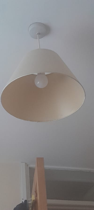 A cream-colored pendant light hangs from a white ceiling fixture. The light has a wide, conical shade with a cream-colored exterior and a lighter cream interior. A single, round, white light bulb is visible inside the shade. Below the light fixture, the upper portion of what appears to be a wooden structure, possibly part of furniture or a ladder, is visible against a pale grey background. There is a white vertical object to the left of the wooden structure, with a yellow tag attached. The scene is indoors and appears to be a domestic environment. There is no discernible evidence to place this scene in Monaghan, Ireland.