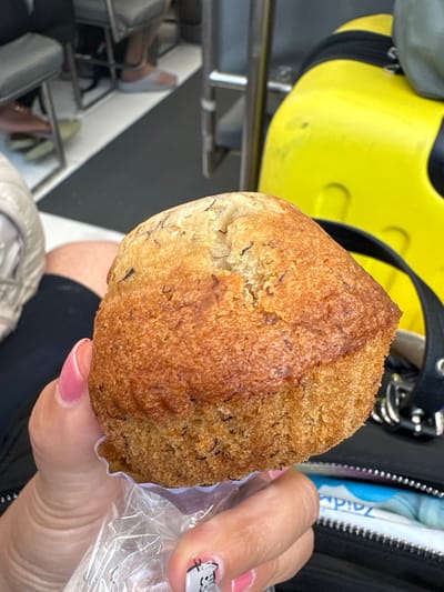 The image captures a close-up view of a banana muffin held by a human hand within an enclosed public setting in Pak Nam, Thailand.
**Subject:** A single, dome-shaped banana muffin with a golden-brown, textured crust and visible dark flecks, indicative of mashed banana or spices, occupies the central foreground. It is partially enclosed in a clear plastic bag and rests on a white, pleated paper liner.
**People & Action:** A human hand, with fingernails painted light pink, holds the muffin from below, gripping the plastic bag. The thumbnail displays a small, light-colored graphic, possibly a cartoon character. In the blurred background, the lower legs and feet of two other individuals are partially visible, one foot wearing a light grey slipper.
**Objects & Environment:** In the lower right background, a black bag or purse with silver-colored metallic rings and a zipper pull is partially visible. Inside this bag, a white and blue packet, possibly containing wet wipes, with partial text "Zaid..." is discernible. To the upper right, a bright yellow, contoured object with black straps, suggestive of a safety floatation device or a piece of luggage, is present. The background also reveals metal structural elements consistent with seating and a floor composed of dark grey or black material bisected by a distinct diagonal white or light grey stripe. This environment suggests the interior of a public conveyance or waiting area, such as a ferry, bus, or train, operating within Pak Nam, Thailand.