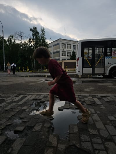 The image shows a young child in a dark red shirt and shorts, wearing light brown sandals. The child is in motion, with one leg extended forward, appearing to be jumping or splashing in a puddle on a cobblestone path. The scene is outdoors, with a cloudy sky suggesting overcast weather, possibly after rain.

In the background, a white bus is parked, with its doors open and yellow handrails visible. Beyond the bus, a multi-story building with many windows stands. Trees line the area behind the building. Several yellow scooters are parked in a row further down the path. Two adults are visible walking in the distance, one wearing a white shirt and dark pants, and another with blonde hair and pink pants.

The overall impression is one of a casual moment of play, possibly in an urban or suburban setting in Novoivanovskoe, Russia. The lighting is dim, indicating it might be late afternoon or early evening, or simply a very cloudy day. There is some text visible on the bus, including "27K".