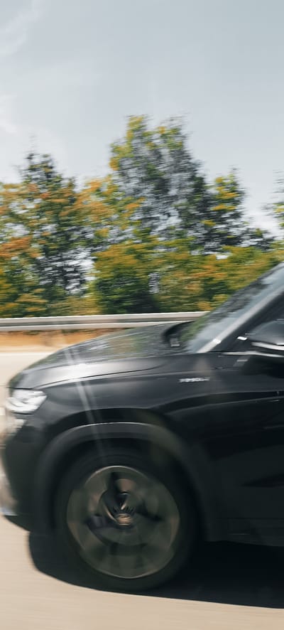 The image depicts a motion-blurred view of the side of a dark-colored car, likely an SUV, traveling on a road. The car's front fender, wheel, and a portion of its hood are visible. The wheel is spinning rapidly, indicated by the blur. In the background, trees with green and yellow foliage are also blurred, suggesting movement and speed. A white guardrail runs parallel to the road in the mid-ground. The sky is overcast and a pale gray. The image does not contain any identifiable landmarks that would specifically place it in Korb, Germany.