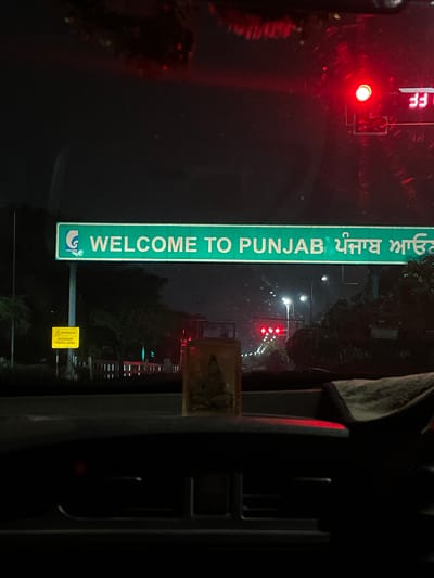 The image captures a nighttime view from inside a vehicle, looking forward. Dominating the scene is a large green overhead sign with white lettering that reads "WELCOME TO PUNJAB" in English, followed by text in Punjabi script. A logo for "GMADA" is visible on the left side of the sign. To the left of the sign, a yellow warning sign with a black border and symbols is attached to a pole. The environment is dark, indicating nighttime, with streetlights and traffic signals visible in the distance, including a red traffic light. Trees and foliage line the roadside. The interior of the vehicle is visible in the foreground, including a dashboard and a small religious figurine placed on it. The overall scene suggests the vehicle is at or near a border crossing into Punjab, possibly on a highway or major road leading into or out of Mohali, India.