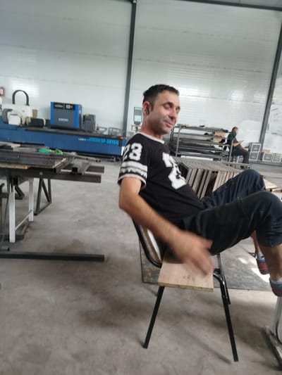 In a large, brightly lit industrial workshop with a concrete floor and white paneled walls, a man wearing a black t-shirt with white lettering and dark pants sits on a simple metal-framed chair with a wooden seat. He has short dark hair and appears to be wearing earbuds. His right arm is in motion, suggesting he might be talking or gesturing. In the background, other individuals are visible, including one person seated and looking at a phone. Various pieces of industrial equipment, such as a blue laser cutting machine labeled "XINFENG", and stacks of metal materials are present throughout the workshop. The scene suggests a working environment, possibly a manufacturing or fabrication facility.