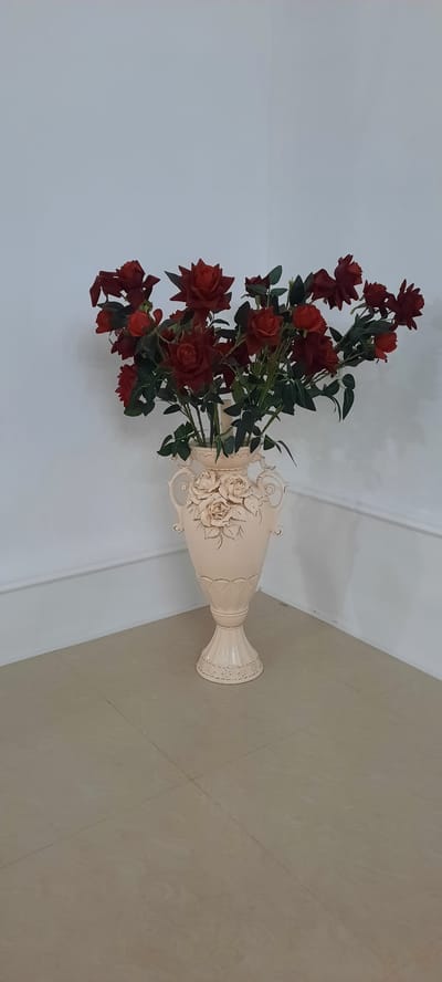 A bouquet of artificial red roses is displayed in an ornate, cream-colored ceramic vase. The vase has a wide opening and is decorated with raised floral patterns and scrolling handles. The roses are a deep red, with some appearing more open and others tightly budded, interspersed with green artificial leaves. The arrangement is positioned in a room with a white wall and a light-colored tiled floor. The tiles have a subtle pattern resembling wood grain and are laid out in a grid with visible grout lines. The scene appears to be indoors, with artificial lighting. There is no indication of the time of day or weather. The location context is Gudermes, Russia, but no specific cues within the image support this. There is no text visible in the image. The overall impression is one of a decorative still life, possibly in a home or a commercial space like a hotel lobby or shop. The lighting is soft and even, suggesting an interior environment.