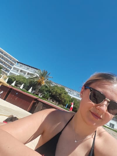 A woman wearing a black bikini and sunglasses is taking a selfie on the beach in Sant Agustí, Spain. In the background, there is a white hotel building with a palm tree and green foliage in front of it. Beach huts are visible on the left side of the image. The sky is clear and blue.