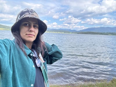 A medium shot portrait of a woman wearing a camouflage bucket hat and a teal corduroy jacket, standing near a lake. She is looking towards the camera with a neutral expression. Her dark hair is visible beneath the hat. A pair of glasses are hanging from the collar of her jacket. The background shows a large body of water, with a shoreline of grass and small yellow flowers. Beyond the lake, rolling hills and mountains are visible under a cloudy sky. The scene is consistent with the natural scenery found in Tsigov Chark, Bulgaria, a resort area known for its lake and mountainous surroundings.