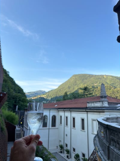 A hand holds a champagne flute filled with bubbly champagne. The glass is in the foreground, with a blurred white building and a lush green mountainside behind it. The building has a red tile roof and arched windows. A stone structure with a spire sits atop the building. The sky is clear and blue, with a few wispy clouds. The sun is setting, casting a warm glow on the scene. The image evokes a sense of relaxation and celebration, with the champagne and the beautiful scenery suggesting a vacation or special occasion. The location appears to be San Pellegrino Terme, Italy, known for its natural beauty and thermal spas.