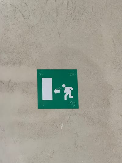 The image shows a green square sign with white pictograms on a textured, light-colored wall. The sign depicts a white rectangle on the left, representing a doorway, with an arrow pointing from the doorway towards a white figure on the right. The figure is stylized and appears to be running or moving away from the doorway. This is a standard exit sign, indicating a place of egress. The wall has an uneven, stucco-like finish with some smudges or dirt marks visible. The lighting suggests an indoor environment. The sign is positioned centrally in the frame. There are no people visible in the image other than the pictogram. The location is specified as Piacenza, Italy.