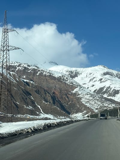 The image captures a mountainous environment in Kanobi, Georgia, under a clear blue sky with a prominent white cumulus cloud in the upper-left quadrant. A paved asphalt road, dark in color and appearing dry, runs from the lower foreground, curving to the right and receding into the mid-distance. Banks of dirty, partially melted snow line the immediate edges of the road, particularly on the left. A tall, dark metal lattice-work electricity transmission tower stands on the far left, extending vertically from the bottom of the frame, supporting three visible power lines that traverse horizontally across the mid-air. In the distance, on the road, a single dark-colored SUV or truck is observed traveling away from the viewer. The background consists of rugged, snow-capped mountains. The lower and middle sections of the mountains display dark brown and reddish rock faces, with varying degrees of patchy snow cover. A small cluster of structures, including a distinct light-colored building with a peaked roof, is visible at the base of the mountains in the mid-left. No individuals are discernible.