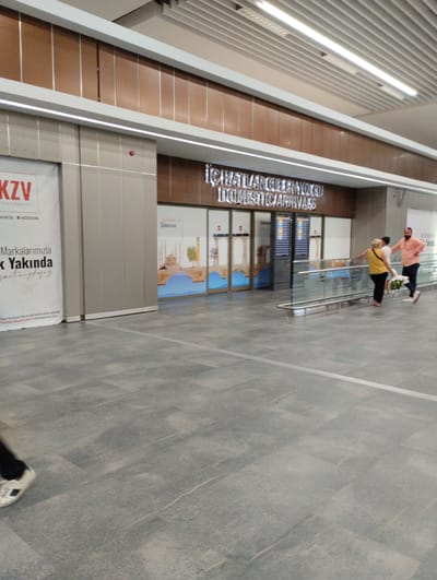 The image captures an interior view of an airport terminal, likely in Tarsus, Turkey, based on the text. The scene depicts a modern, well-lit space with polished gray tiled flooring. In the background, a sign prominently displays "İÇ HATLAR / DOMESTIC ARRIVALS" in large, metallic letters, indicating this is the domestic arrivals section of the airport.

Several people are visible. In the center-right of the image, a woman wearing a yellow top and black pants is walking with a young boy. Behind them, a man in a peach-colored shirt and dark pants is walking away from the camera. The man appears to be holding a bouquet of white flowers, suggesting he might be picking someone up or returning home with them.

To the left, a large banner with red and white branding reads "KZV" and mentions "Havalimanları" (Airports). Further text on the banner indicates "Markalarımızla Yakında" (Soon with our brands) and a website or email address "icarinsler@kozuva.com.tr". The overall atmosphere appears to be that of a typical airport arrival hall, with people moving through the space. The lighting suggests it is daytime.