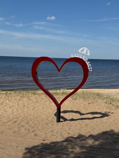 An outdoor scene captures a large red heart-shaped sculpture on a sandy beach, overlooking a vast expanse of water under a clear blue sky. The image is taken on a bright, sunny day, evidenced by the distinct shadows cast on the sand, suggesting either early morning or late afternoon.
The prominent feature is a red, stylized heart sculpture, positioned on the light-colored sand. Attached to the upper left curve of the heart is a white metal sign. This sign features an umbrella icon and the word "SAULKRASTI" in capital letters, oriented vertically. This text, combined with the location context, confirms the setting as Saulkrasti, Latvia.
Behind the heart sculpture, the calm waters of the sea (likely the Baltic Sea) stretch horizontally across the frame, meeting a clear sky with a few wispy clouds. The water exhibits subtle ripples.
In the foreground and to the right of the heart, a wide sandy beach is visible, with patches of sparse green grass growing along the edge where the sand meets the water. Long, dark shadows are cast across the sand; one distinct shadow appears to be that of a person standing, while other, more amorphous shadows suggest additional objects or foliage just out of frame. The sand shows gentle undulations and scattered small debris. The overall impression is one of a peaceful, scenic coastal location.