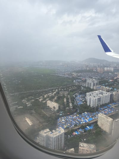 An aerial view from an airplane window shows the cityscape of Vindhane, India. The image captures a dense urban environment interspersed with green spaces. In the foreground, the curved edge of the airplane window frames the scene. A portion of the airplane's wing, predominantly white with a blue tip, is visible in the upper right corner.

The landscape below reveals a mixture of housing types. There are numerous tightly packed, low-rise structures, many of which are covered with blue tarpaulins, suggesting a densely populated residential area, possibly a slum or informal settlement. These are contrasted with several modern, multi-story apartment buildings and office blocks, characterized by their rectangular shapes and light-colored facades. A railway track runs through the settlement, with a train visible on it. A narrow waterway or canal also winds through the urban area.

Further into the distance, large tracts of green land are visible, possibly agricultural fields or undeveloped areas. Beyond that, the city extends into a hazy horizon, with more buildings and the outline of hills or mountains in the far background. The sky is overcast, with a heavy cloud cover that creates a muted, diffused light across the entire scene, indicating a possibly rainy or foggy day. The overall impression is one of a sprawling city with significant contrasts in development and density.
