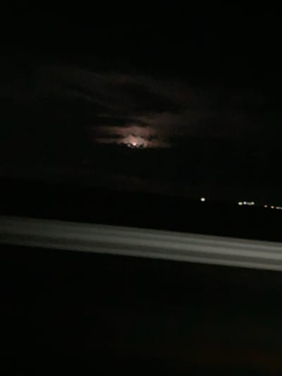 A nighttime photograph captures a dimly lit scene. The moon is visible through scattered clouds in the upper central portion of the image, casting a faint glow. Below the moon, the dark expanse of the sky stretches across the frame. In the distance, several blurred lights suggest the presence of a distant urban area or coastline, possibly in the city of Damsa, Kazakhstan, given the context. In the foreground, a horizontal, light-colored stripe, possibly a railing or a car's interior, bisects the lower half of the image, suggesting the photograph was taken from a moving vehicle or a similar vantage point. The overall impression is one of a quiet, nocturnal landscape with the moon as the primary celestial feature.