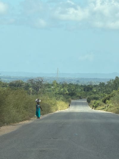 The image shows a rural scene, likely near Muxara, Mozambique. An asphalt road curves to the right of the frame. Green vegetation lines the roadside. A person wearing a blue dress and a white head covering stands beside the road. A distant landscape of trees and possibly mountains fades into the background under a blue sky with clouds. A utility tower is visible in the mid-distance.