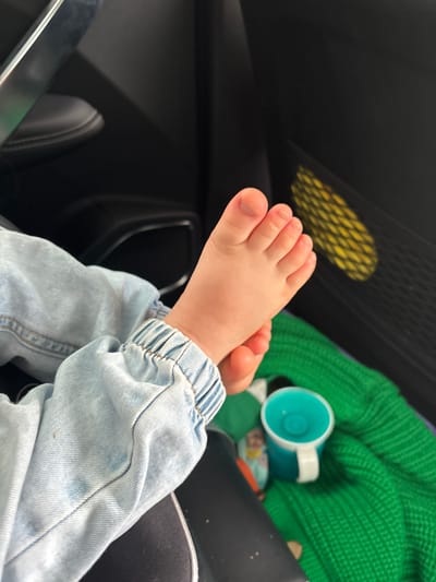 The image shows a close-up of a baby's feet, wearing light blue denim pants with elastic at the ankle, resting on a green knitted blanket. A turquoise sippy cup with a white handle is also visible on the blanket. In the background, dark car interior elements, including a seat and a mesh storage pocket with a yellow object inside, are discernible. The lighting suggests an indoor environment, possibly a car. The scene is devoid of any specific indications of the city of Боровляна, Russia.