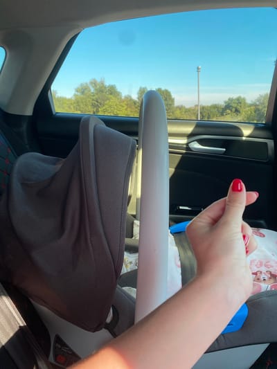 A person's hand with red nail polish is making a finger heart gesture in the foreground of a car interior. The hand is positioned in the right side of the frame, obscuring part of a car seat and a white cylindrical object. Behind the hand and the object, a dark gray car seat is visible, covered with a patterned fabric in some areas. Through the car window in the background, a clear blue sky is visible, with a line of green trees and a lamppost. The scene is lit by natural sunlight.