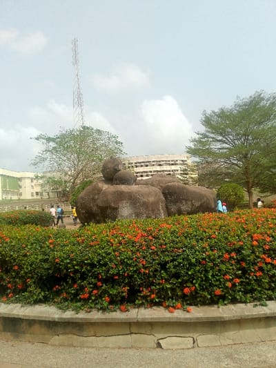 This image depicts an outdoor scene in the city of Adzho, Nigeria. In the foreground, a low, cracked concrete wall forms the base of a planter filled with dense green bushes adorned with numerous small, clustered orange-red flowers.

Dominating the mid-ground is a cluster of large, smooth, dark grey boulders, some appearing stacked. To the left of these boulders, several individuals are visible walking on what appears to be a paved area. One individual wears a pink shirt and blue trousers, another has a blue backpack, and a third wears a yellow top. To the right of the boulders, a person in light blue attire with a red head covering is partially visible behind the flowering bushes.

In the background, a light-colored multi-story building with visible windows and green accents is present on the far left. Towards the center-right, another multi-story building with horizontal window bands is visible. Several trees with green foliage are scattered across the mid and background, some partially obscuring the buildings. A tall, slender lattice communications tower extends vertically into the sky from behind a tree on the left side of the frame. The sky is overcast with scattered white and light grey clouds.