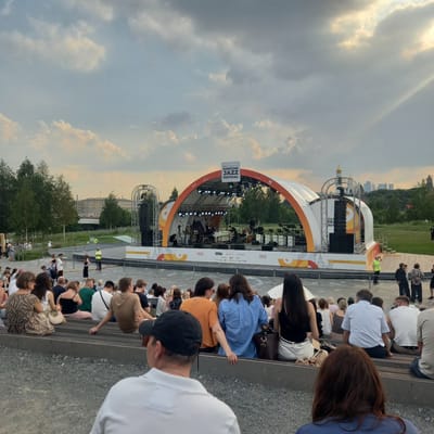 The image depicts an outdoor concert venue with a stage and an audience seated on tiered benches. The stage is large and arched, decorated with the words "MOSCOW JAZZ FESTIVAL" and sponsors' logos. A band is performing on stage, with musicians playing instruments and equipment such as speakers and lighting rigs visible. The audience is composed of a diverse group of people, many of whom are seated, facing the stage. Some individuals are standing or walking in the background. The environment appears to be a park or open space, with trees and grassy areas surrounding the seating area. The sky is cloudy, with some sunlight breaking through. The context of "Pутрышкаўскі сельскі Савет, Belarus" is not evident in the visual information provided by the image itself.