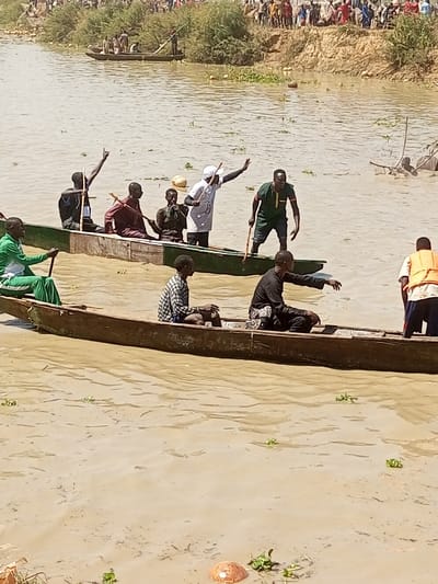 The image depicts a crowded water scene in Kokani North, Nigeria, featuring multiple individuals in wooden dugout canoes on murky brown water.

In the foreground, two long, narrow wooden dugout canoes occupy the center. The lower boat, dark brown, carries three males. From right to left: one male stands, facing away from the viewer, wearing a white and orange life vest. Adjacent to him, a male in dark clothing sits facing left, extending his right arm forward. To his left, another male in a plaid shirt and shorts sits facing left.

The upper boat, painted green, carries five males. From right to left: one male stands, wearing a dark green shirt and shorts, holding a wooden pole, and looking toward the left side of his boat. Next to him, a male in a white long-sleeved shirt and white cap stands, holding a pole, with his left arm raised and extended. To his left, a male in dark clothing is seated, holding a pole. Further left, a male in a reddish-brown garment is seated, holding a pole, with his left arm raised and hand pointing upward. Furthest left, a male in a green tracksuit sits, holding a paddle or pole, facing left.

Between the two main boats, a white spherical buoy floats on the water. The water itself is a light brown, indicative of suspended sediment, with sparse green aquatic vegetation visible. A half coconut shell and other debris float in the lower portion of the image.

In the mid-ground and background, a sandy, elevated bank is visible on the far side of the water, topped with dense green shrubbery and trees. A large crowd of people is gathered on this bank, observing the scene in the water. Another smaller, dark-colored dugout canoe is visible further upriver, carrying 3-4 individuals, with one standing and holding a pole. To the right of this distant boat, a partially submerged object or individual is visible in the water. The sky is bright and clear, suggesting daylight conditions.