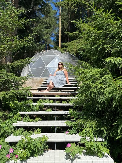 In Makarovo, Russia, a woman sits on the steps of a concrete staircase outdoors. She is wearing sunglasses and a blue and white patterned dress. The staircase is surrounded by lush green trees and vegetation, and behind it, a geodesic dome structure is visible. The sunlight suggests it is daytime.