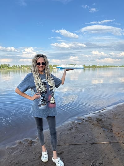 A woman with long blond hair is standing on a sandy beach at the water's edge in Sosnovtsy, Russia. She is wearing dark sunglasses, a gray t-shirt with a print, gray jeans, and white Crocs. She has one hand on her hip and the other raised in the air. Behind her is a body of water with a cruise ship on the left and a tall structure in the distance, indicating an industrial area. The sky is blue with white clouds. The photo has been taken from a rotated angle.
