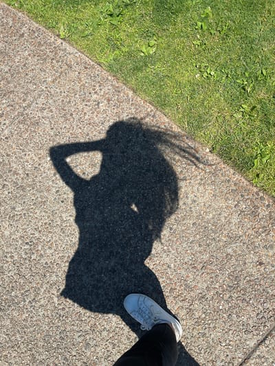 The image is a high-angle shot of a shadow on a concrete path next to a patch of grass. The shadow appears to be of a person with long hair, seen from the waist up, with their arm raised and hand on their head. A white sneaker and black pants are visible in the bottom right corner, belonging to the person casting the shadow. The concrete path is made of small, mixed-colored pebbles. The grass is green and appears to be well-maintained. The lighting suggests it is daytime, with strong sunlight casting a distinct shadow. The location is the town of Oak Bluffs, United States of America.