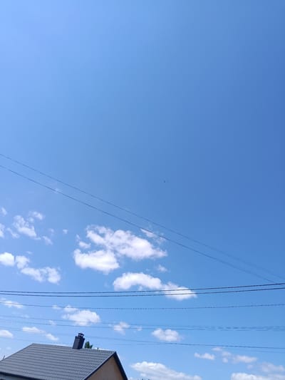 The image captures a clear, bright blue sky with a few scattered white clouds. Several power lines crisscross the frame horizontally at different heights. In the lower portion of the image, the roofline of a grey-tiled building is visible, featuring a dark-colored chimney. The scene appears to be an outdoor environment, likely residential or suburban, in Kostopil, Ukraine. There are no people or animals clearly discernible in the image. A faint, distant object, possibly a bird or aircraft, is present in the upper central sky.