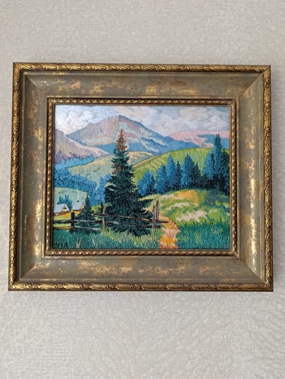 A framed needlepoint artwork hangs on a textured, off-white wall. The artwork depicts a mountainous landscape, possibly a scene in or reminiscent of the Ukrainian countryside. In the foreground, a rustic wooden fence stretches across a field of tall grass and wildflowers. Behind the fence stands a large evergreen tree. To the left, a small yellow building can be seen nestled among other trees. In the background, rolling hills and mountains fade into a soft blue and pink sky. The artwork is encased in an ornate gold and green frame. There is no evidence to confirm the location within the city of Barvinok, Ukraine.
