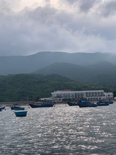 The image depicts a serene coastal scene with a backdrop of lush, green mountains and a building complex. The foreground is dominated by the sparkling, sunlit surface of the water, suggesting a bright and sunny day. Several blue boats are moored along the shore, indicating a fishing or leisure activity. The building complex appears to be a hotel or resort, with its multi-story structure and numerous windows. The mountains in the background are shrouded in a light mist, adding a sense of depth and tranquility to the scene. The overall mood is peaceful and picturesque, evoking a sense of calm and relaxation. The time of day appears to be late afternoon or early evening, given the angle of the sunlight reflecting on the water. There is no visible text in the image.