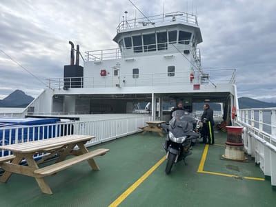 A ferry deck is shown under an overcast sky. Two men in motorcycle gear stand next to two motorcycles. The men are wearing black jackets and pants, and one man has bright yellow stripes on his pants. The motorcycles are parked on the green deck, which has a yellow line painted on it. A picnic table is to the left of the frame. In the background, a large white building with windows is visible, which appears to be the ferry's superstructure. Mountains are seen in the distance. The scene is likely on a ferry operating in or near Nesna, Norway.
