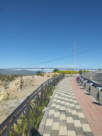 The image shows a paved walkway with a black metal fence on the left, separating it from a rocky, uneven terrain. The walkway itself is paved with alternating light gray and white square tiles, with a section of reddish-brown rectangular tiles on the right edge. A metal guardrail runs along the outer edge of the walkway, adjacent to a paved road. In the background, a long suspension bridge spans a wide canyon, with mountains visible in the distance. A tall, metallic tower stands near the bridge on the right side of the frame. The sky is clear and blue. The location is identified as the city of Myk, Uzbekistan.