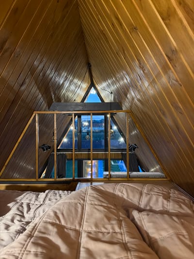 The image shows the interior of a wooden A-frame cabin. The perspective is from a bed, looking up towards the apex of the roof. The walls and ceiling are paneled with light brown wood. In the midground, there is a wooden railing overlooking a lower level with large windows. Through the windows, the exterior landscape can be seen at dusk, with lights from the cabin and possibly the surrounding area visible. There are decorative Edison-style light bulbs hanging from the ceiling. An air conditioning unit is visible on the lower level. The bed in the foreground is covered with a light brown quilted comforter. The overall scene suggests a cozy, rustic accommodation. The image is located in Sharovechka, Ukraine.