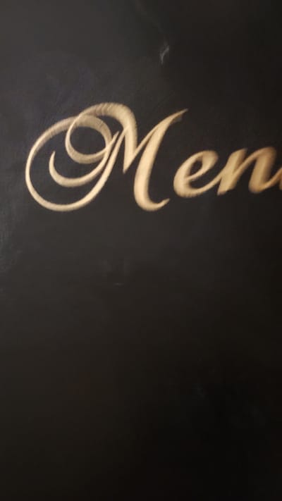 The image shows the word "Menu" written in a cursive, gold-colored font on a black surface. The word is positioned in the upper portion of the image, with the letters appearing to be raised or embossed. The black surface has a textured appearance, and there are some faint, lighter marks visible on it, possibly due to lighting or slight imperfections. The background is entirely dark and featureless. There are no people, actions, or specific environmental details identifiable in this image, nor is there any information about its location in Pevtsite, Bulgaria.