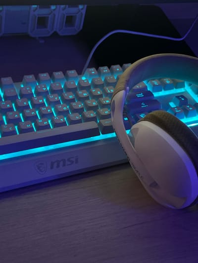 The image displays a close-up, slightly angled view of a white gaming keyboard with blue backlighting, positioned on a wooden desk. The keyboard features Cyrillic keycaps. Partially obscuring the keyboard in the foreground is a white and gray headset with a textured gray earcup. The headset has text printed on its headband, which appears to read "ARENA GAMING TEMPLE". The MSI logo is visible on the front edge of the keyboard. The overall lighting is dim, with the blue glow from the keyboard dominating the scene. The background is dark and out of focus, with what appears to be part of a computer monitor or screen at the top. There are no people present in the image. The scene is indoors, likely a home or gaming setup.