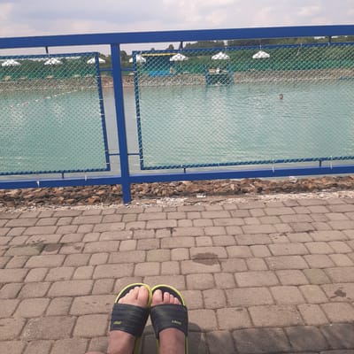 This image shows a close-up of a person's feet wearing black and yellow slide sandals, standing on a paved walkway made of interlocking stone blocks. The walkway is bordered by a gravel area and a blue metal fence with green netting. Beyond the fence is a body of water, possibly a pond or lake, with some white objects floating in it. The water appears a light turquoise color and there are trees visible in the distance on the far side of the water. The scene is outdoors, with daylight suggesting it is daytime. The lighting and shadows indicate a sunny day. The image is taken from a low angle, focusing on the feet and the immediate surroundings. The overall impression is casual and relaxed, as indicated by the footwear and the outdoor setting. The text "SPORT" is visible on the strap of each sandal. The image was taken in Dubrivka, Ukraine.