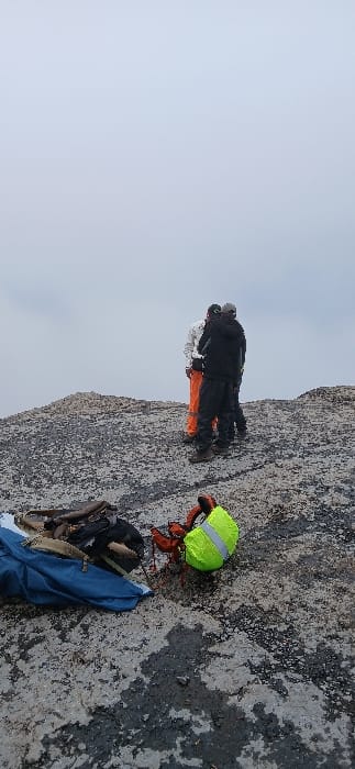 The image depicts a high-altitude, rugged outdoor environment at Gilman's Point, Tanzania, under a uniformly overcast light-gray sky. The ground is an irregular, mottled surface of dark gray rock or earth with lighter gray patches, suggesting dampness or ice.
Three individuals are visible, clustered together towards the upper-right quadrant, facing away from the camera. The leftmost individual wears a white top and bright orange trousers. The central individual wears dark clothing, including a dark jacket and trousers, with a dark cap. The rightmost individual also wears dark clothing and a light-colored cap. All individuals appear to be dressed in cold-weather or trekking attire. Their posture indicates they are standing and observing the vista beyond the frame.
In the left foreground, a blue fabric item, possibly a tent or tarp, is partially visible. Adjacent to it lies a dark backpack with light brown straps, accompanied by several brown, cylindrical objects. In the center foreground, a neon yellow-green backpack, featuring a reflective strip and orange straps or harness, rests on the ground.