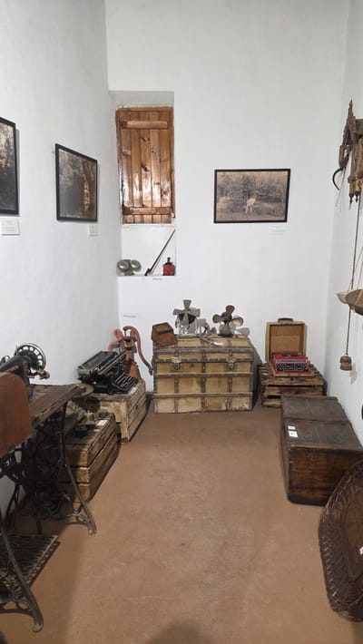 The image captures an interior space in Altura, Spain, likely a museum or historical display. The walls are white. On the walls hang framed black and white pictures, and there is a small wooden shuttered window. A variety of antique objects are scattered on the floor: trunks, a vintage typewriter, old electric fans, a sewing machine, and other miscellaneous items. The floor is a terracotta color. The lighting is ambient and there are no people visible in the image.