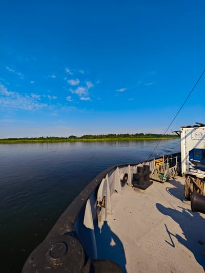 This image depicts a view from the deck of a vessel on a river, likely in or near Novonazimovo, Russia, given the context. The sky is a clear, vibrant blue with sparse, wispy white clouds. To the left of the frame, a calm, dark river stretches towards a distant, tree-lined shore. The shore is a continuous band of green foliage, suggesting a natural landscape.

The foreground is dominated by the deck of the vessel, which is made of a gray, weathered material. Several objects are visible on the deck, including large black bollards, a coiled rope on a spool, and a white shipping container with blue accents and visible lettering that reads "R-134a". Shadows of what appear to be people are cast across the deck, indicating the presence of at least two individuals. One shadow is particularly distinct, showing a figure with an arm raised. A black railing runs along the edge of the deck. The vessel appears to be in motion, with subtle ripples visible on the water's surface. The overall atmosphere is serene and suggests a peaceful journey on the water under a bright sky.