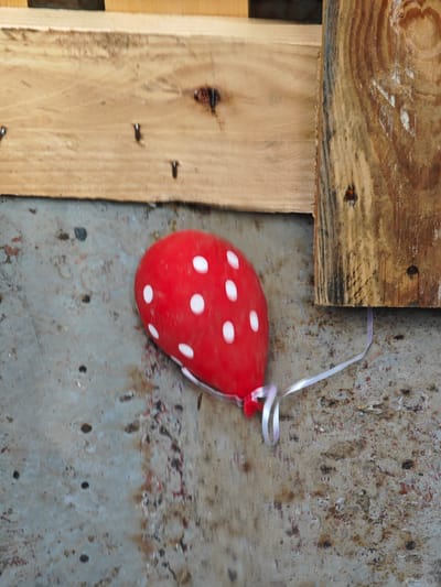 A red balloon with white polka dots is resting on a mottled, grey concrete surface. A thin, white ribbon is tied to the balloon and trails to the right. The background consists of wooden planks. The scene is located outdoors, likely in an urban or semi-urban setting, possibly in Погодаево, Russia. The lighting suggests daytime. There are no people or animals visible in the image. The overall condition of the environment appears somewhat weathered.